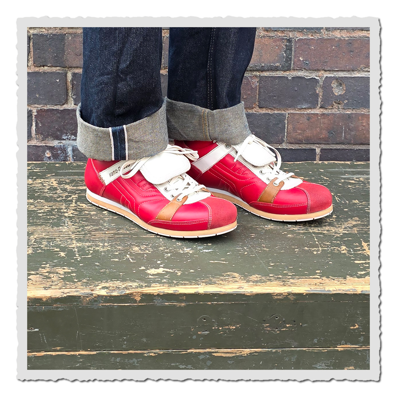 Rote Schnürschuhe mit heller Lasche an einer Person mit dunklen Jeans, vor Backsteinwand auf grünem Holzpodest.